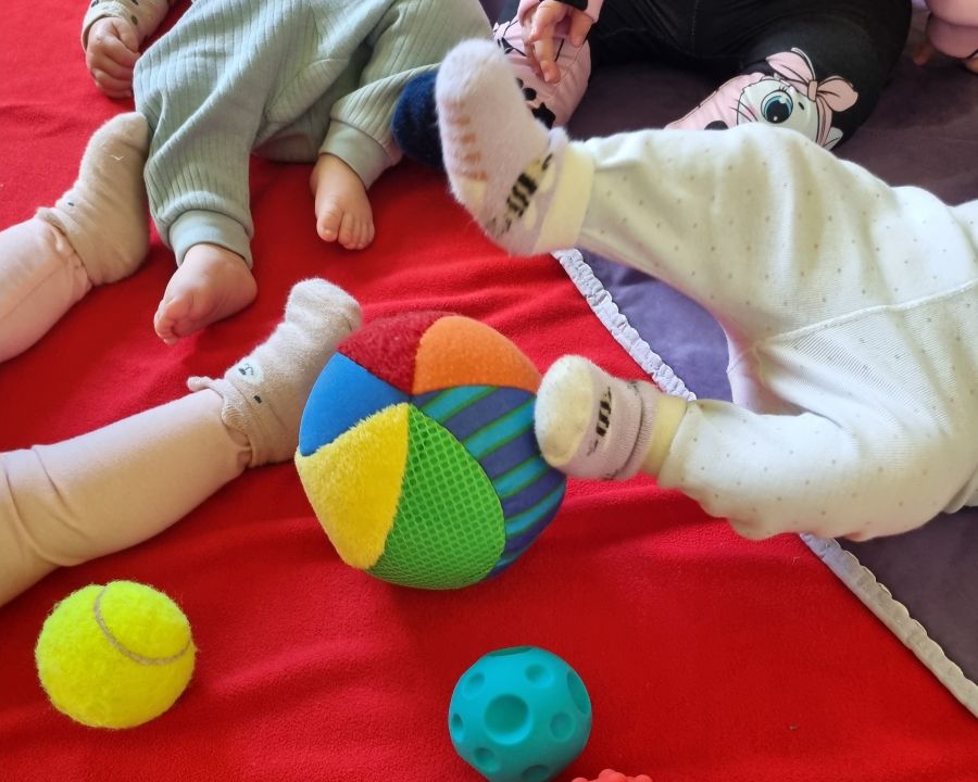 Das Foto zeigt mehrere Bein-Paare von Babys und ein paar bunte Bälle auf einer roten und einer violetten Fließdecke. | Foto: Caritas