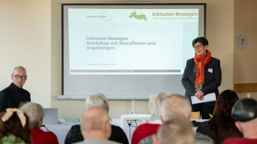 Das Bild zeigt eine Gruppe Menschen vor einer Leinwand, es steht rechts Regina Meyer und begrüßt die Menschen.