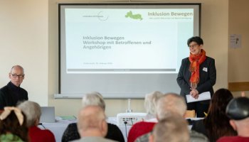 Das Bild zeigt eine Gruppe Menschen vor einer Leinwand, es steht rechts Regina Meyer und begrüßt die Menschen.