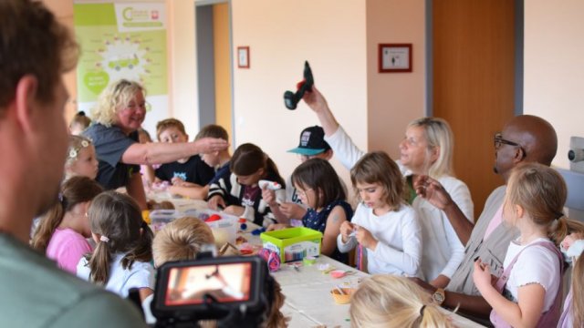 Das Foto zeigt eine Gruppe bastelender Kinder, über die hinweg eine Heißklebepistole weitergereicht wird.
